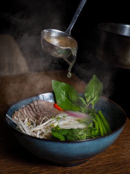 A delicious bowl of pho with steaming broth and fresh vegetables pouring from a ladle.