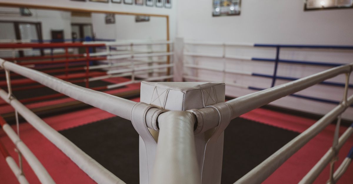 An empty boxing ring inside a gym with red and blue corner ropes and pink flooring.