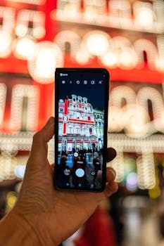 A hand holding a smartphone capturing a brightly lit building at night, showcasing vibrant and colorful city life.