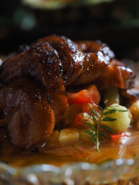 A whole roast duck with deep mahogany, crackling skin resting on a carving board, with a glossy orange sauce being spooned over sliced pieces on a white platter.
