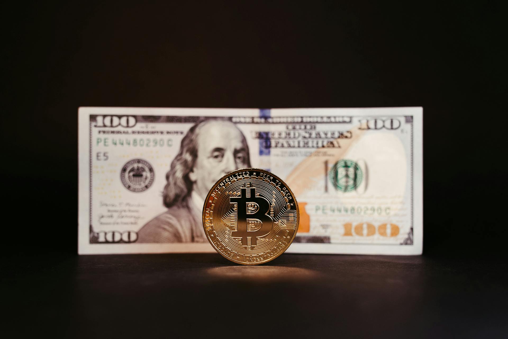 Bitcoin coin standing in front of a 100 dollar bill, symbolizing cryptocurrency and traditional currency themes.