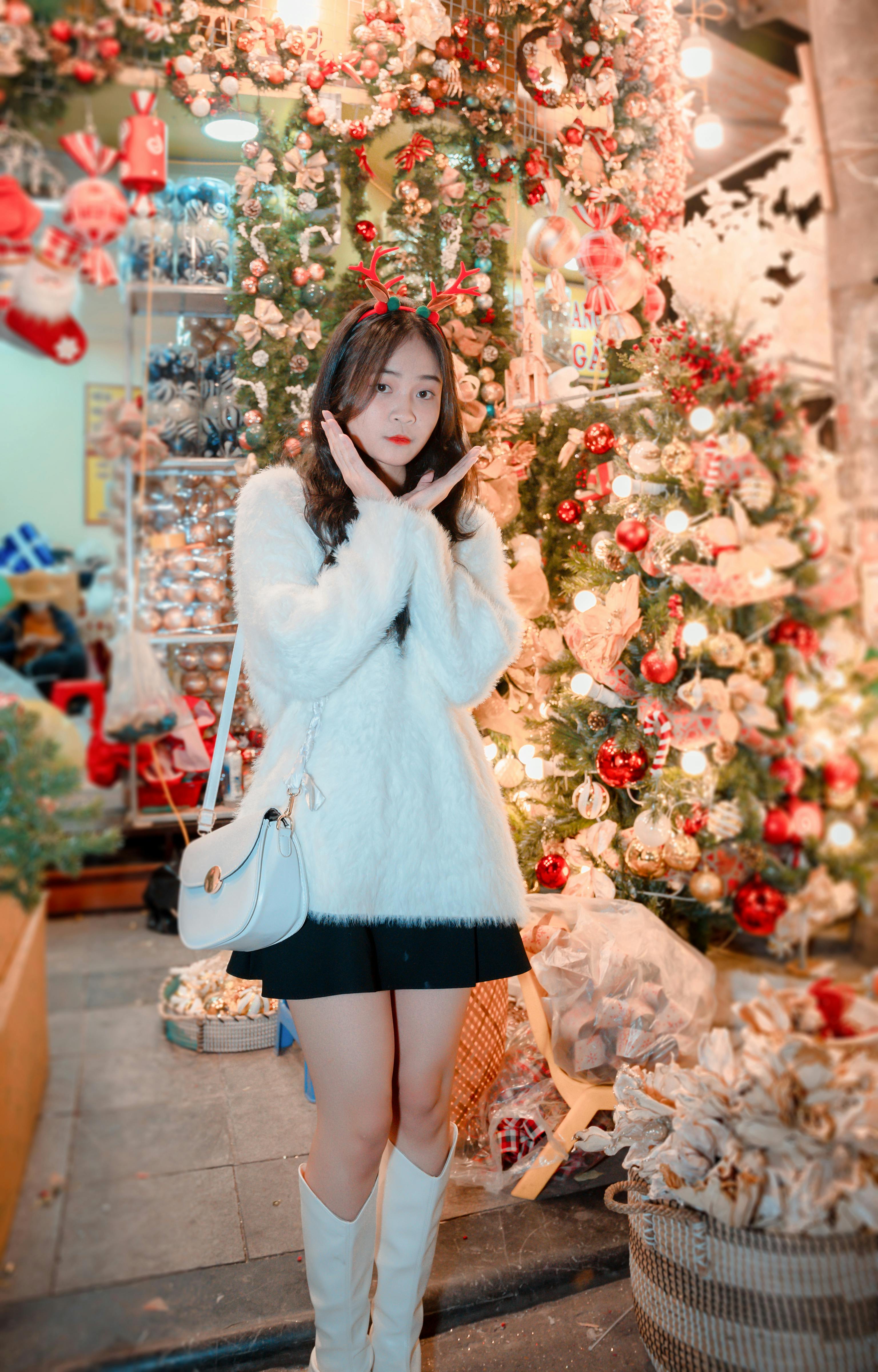 An Asian woman in a white sweater poses by a decorated Christmas tree with ornaments.