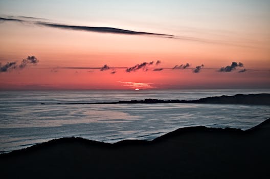 Tranquil sunset over a calm ocean with silhouetted hills and vibrant sky.
