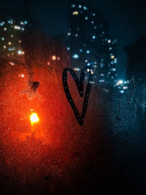Night view of a heart drawn on a steamy window with urban lights in the background.