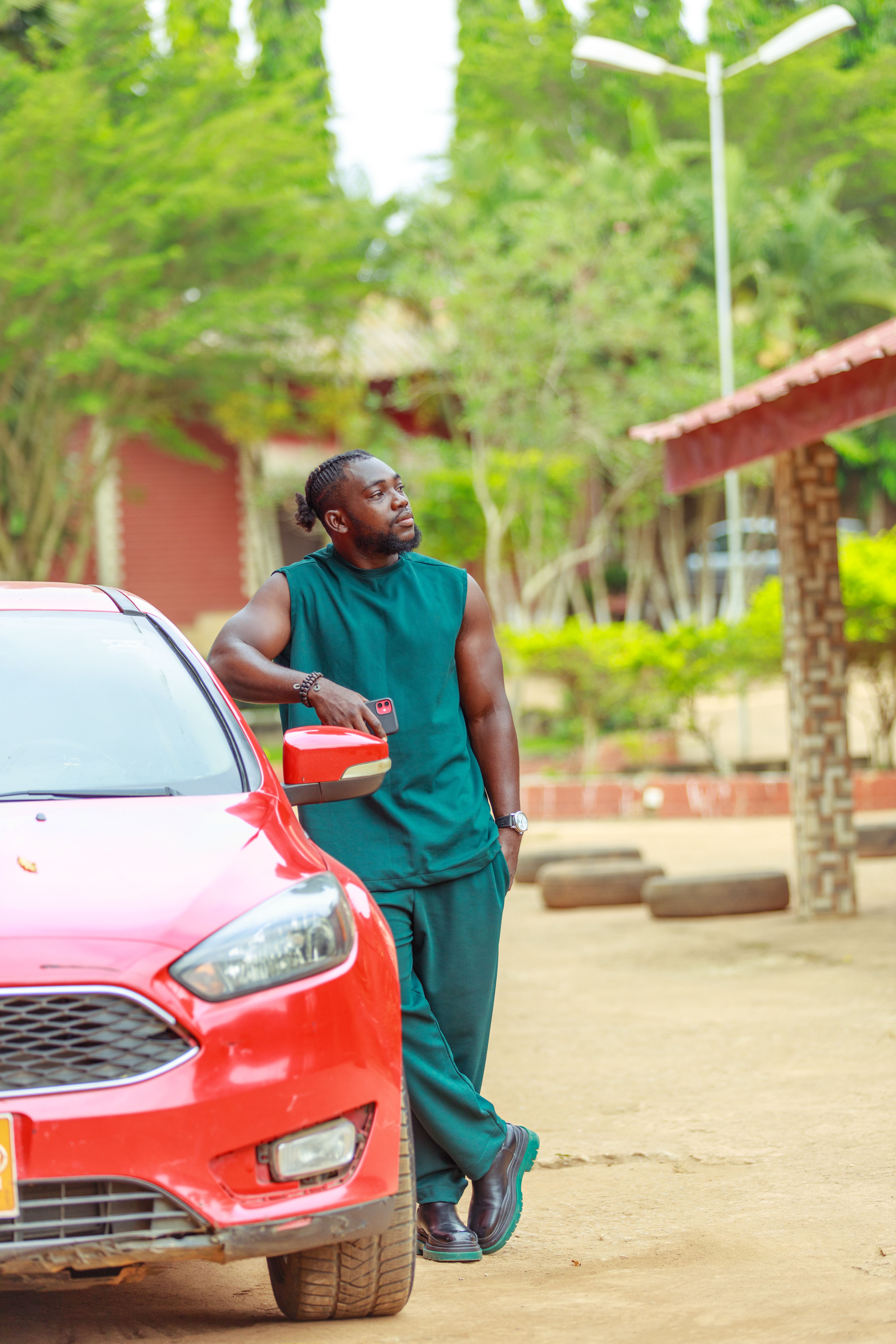 Man in casual fashion leaning on a red car outdoors, using smartphone.