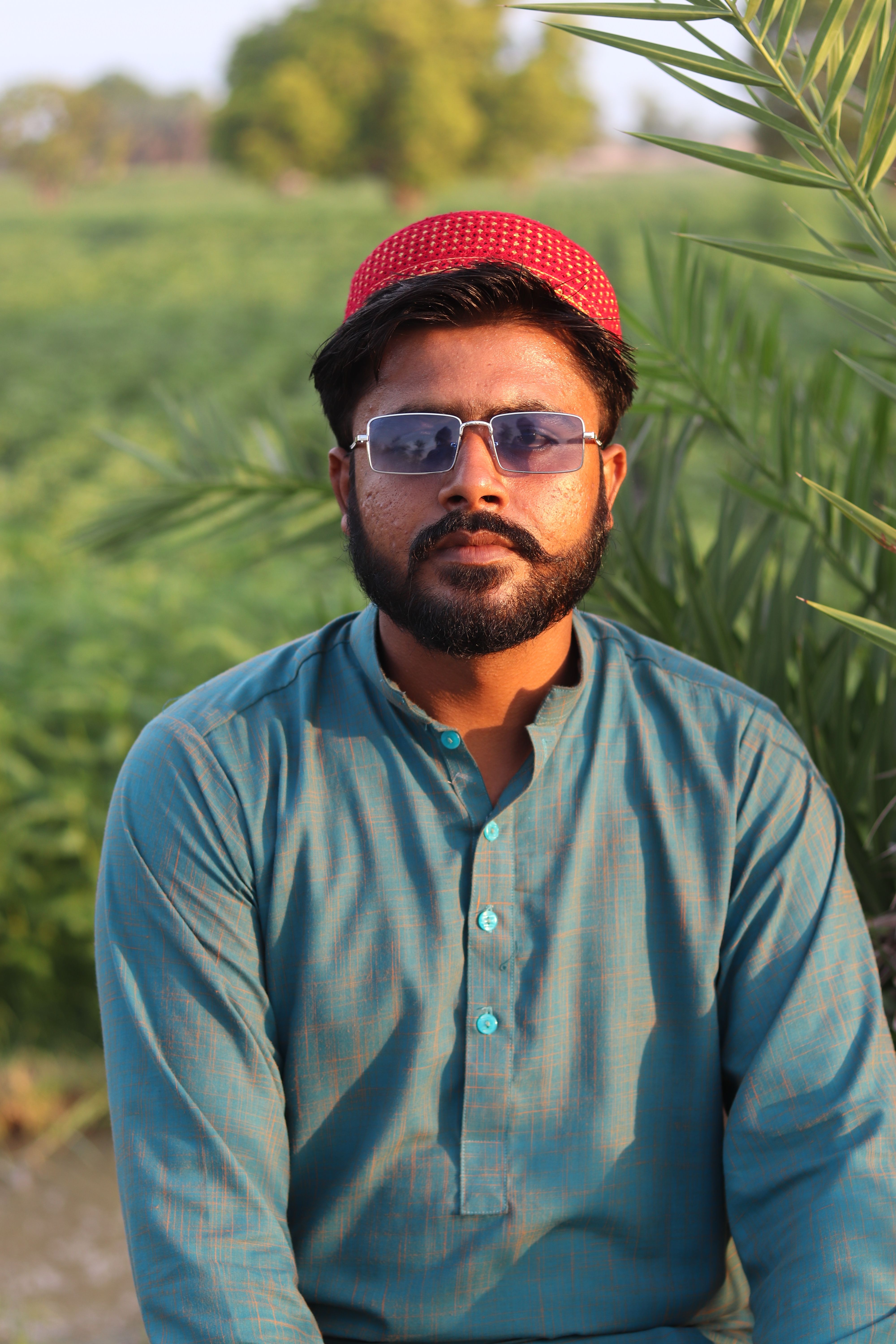 Portrait of a man outdoors wearing a red cap and sunglasses against a green rural background.