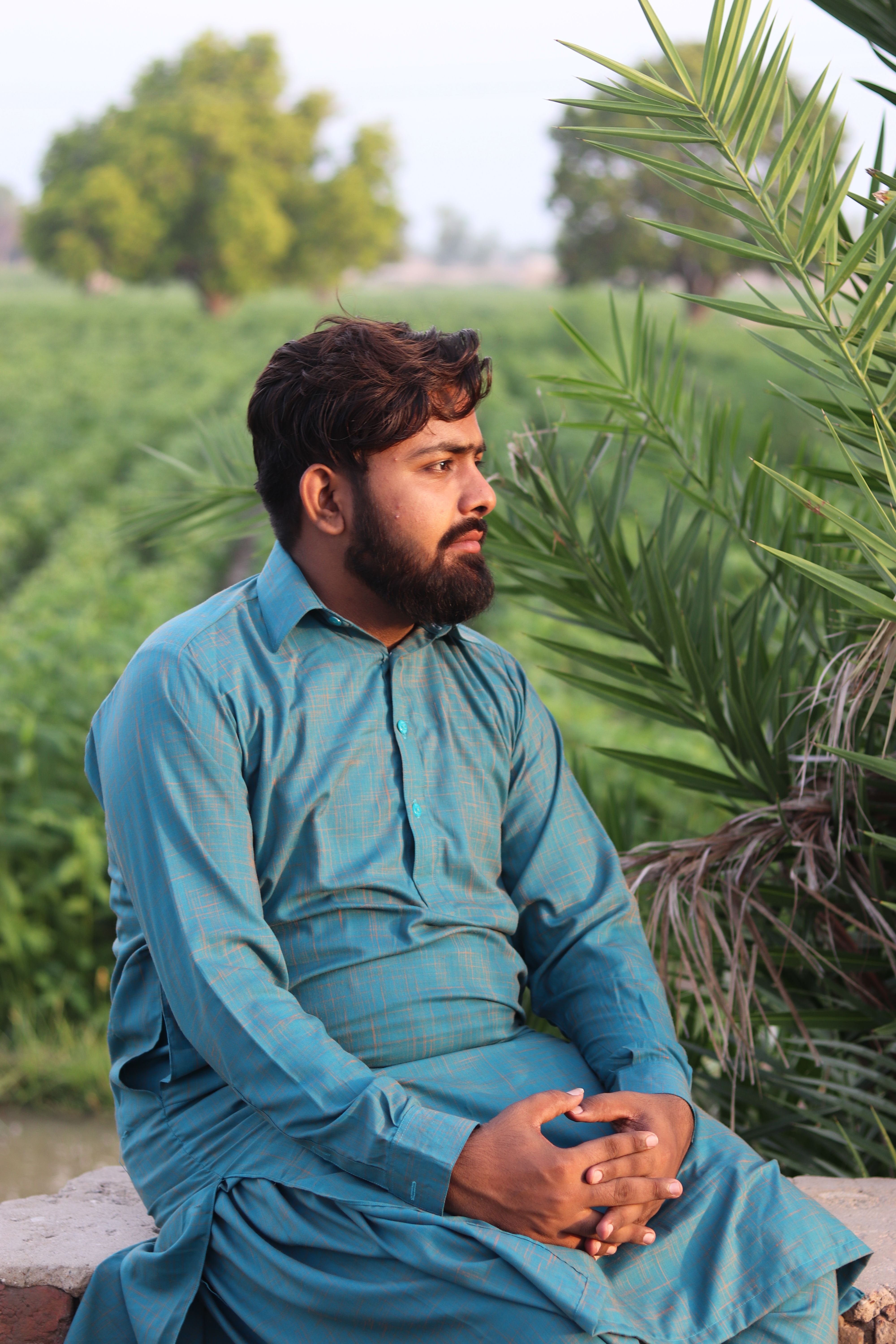 An adult man in traditional clothing sits thoughtfully in a lush, green outdoor setting.