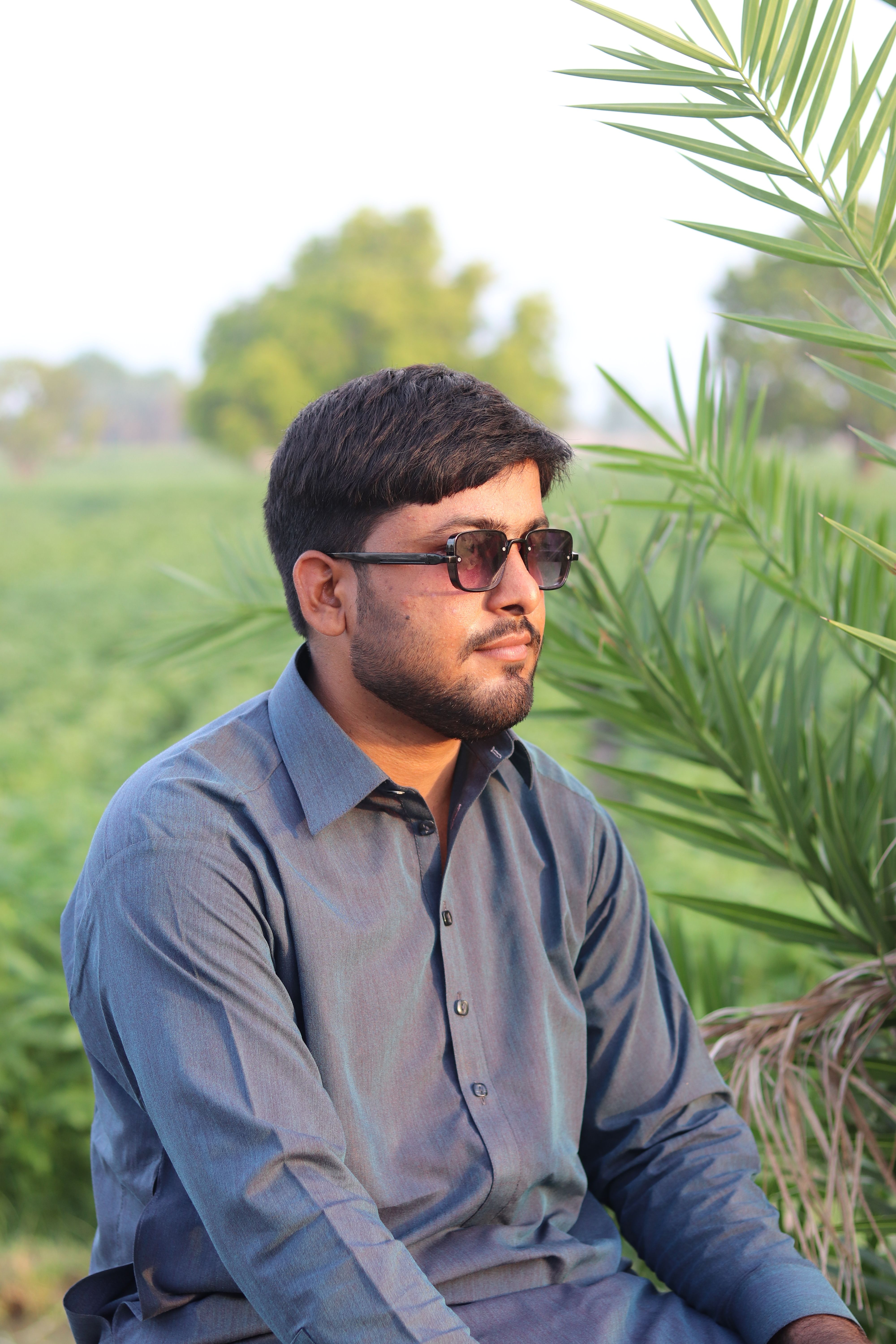 A young man wearing sunglasses sits amidst greenery with a relaxed expression in a lush outdoor environment.