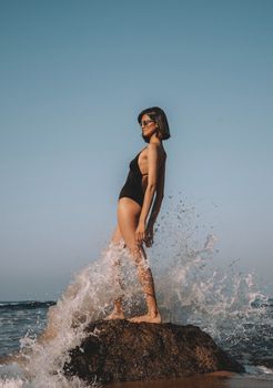 Elegant woman in swimwear posing on rock with splashing waves in Istanbul.