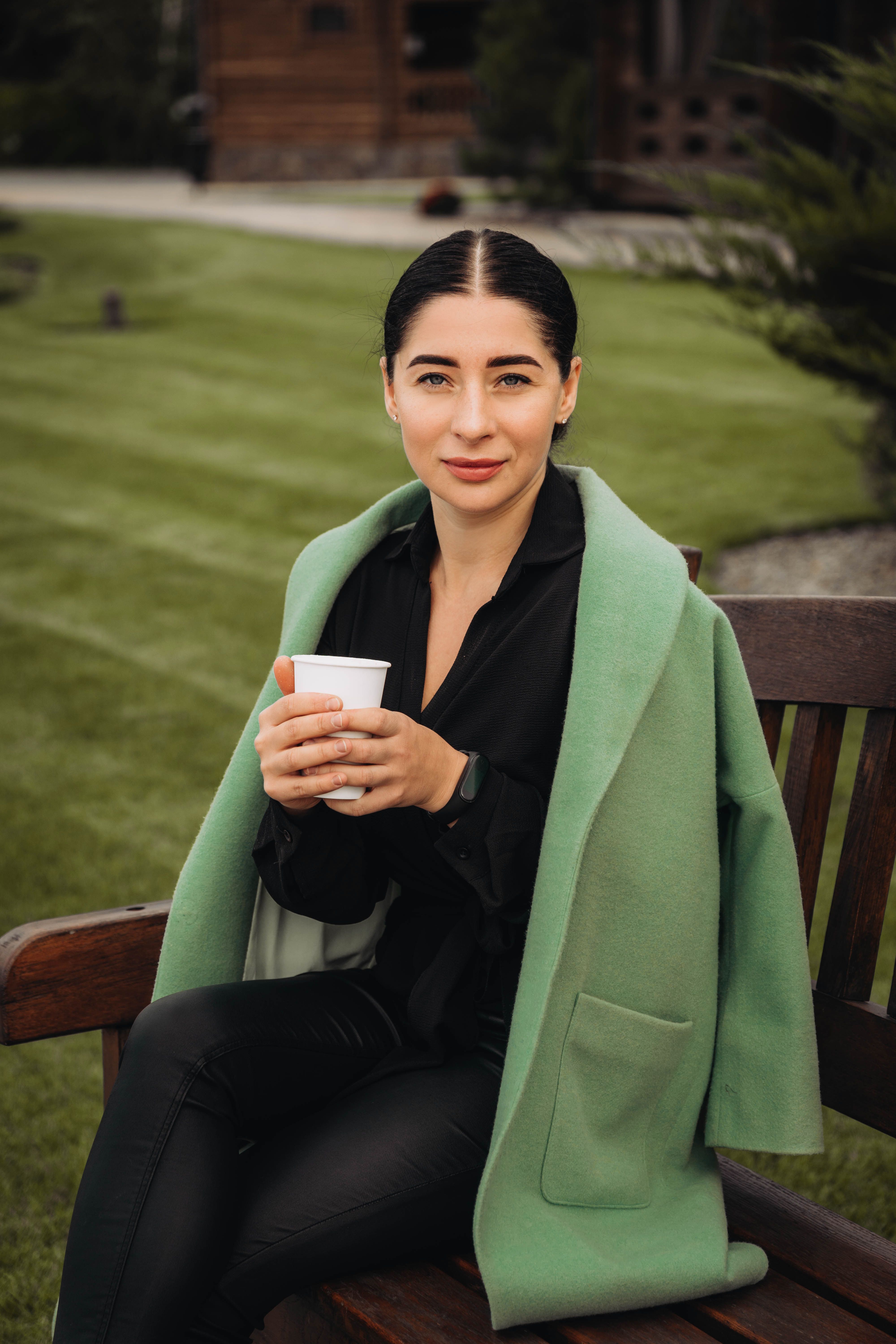 Woman in a stylish green coat enjoys a warm drink on a wooden bench outdoors.
