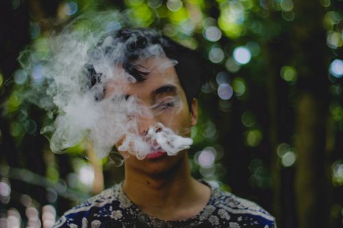 Free A young man smoking and exhaling smoke outdoors with a blurred natural background. Stock Photo
