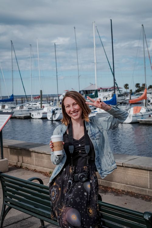 Free A cheerful woman with a camera and coffee making a peace sign by the marina. Sunny day with yachts in the background. Stock Photo