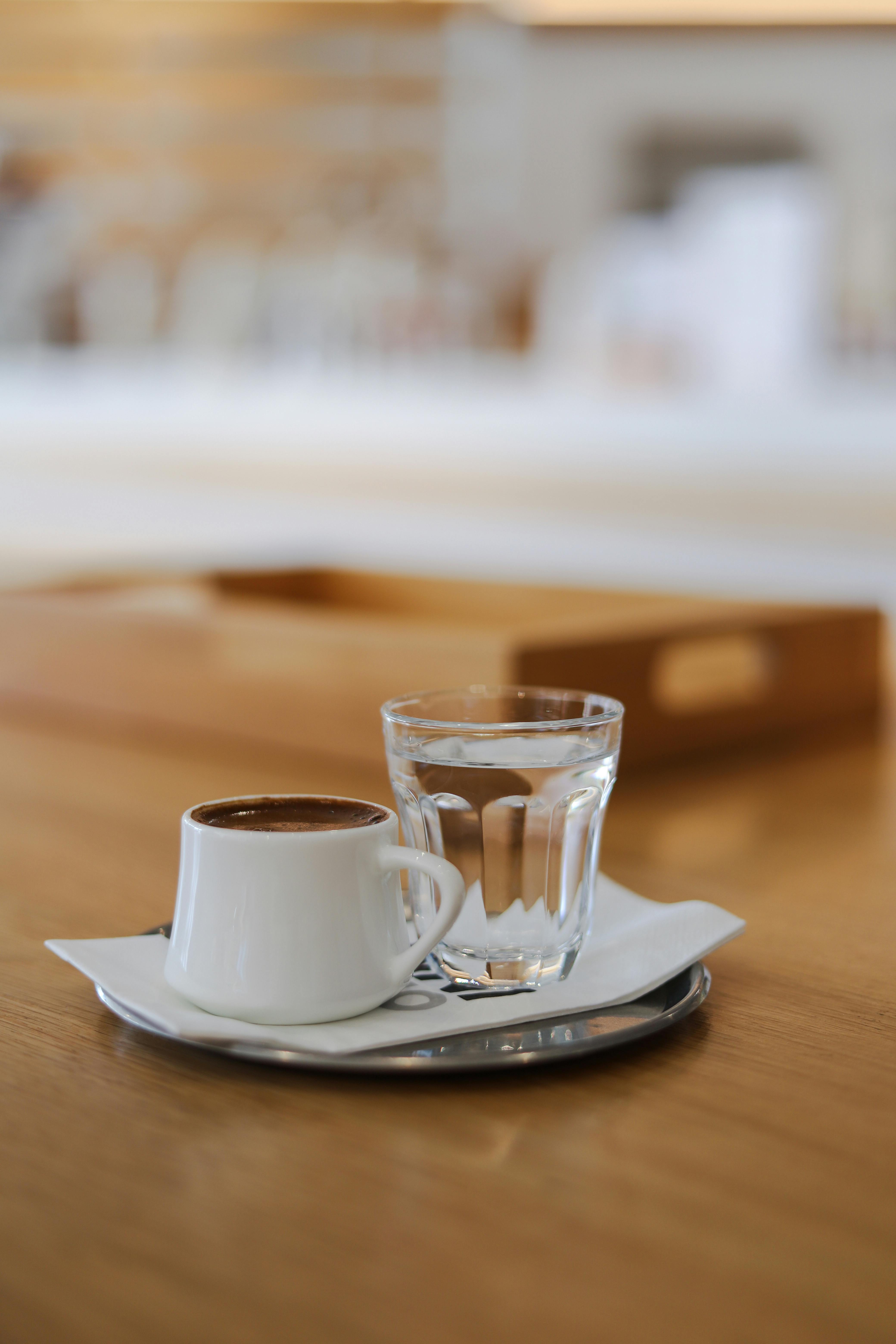 Close-up of a cup of coffee with water on a wooden table indoors, featuring a blurred background.