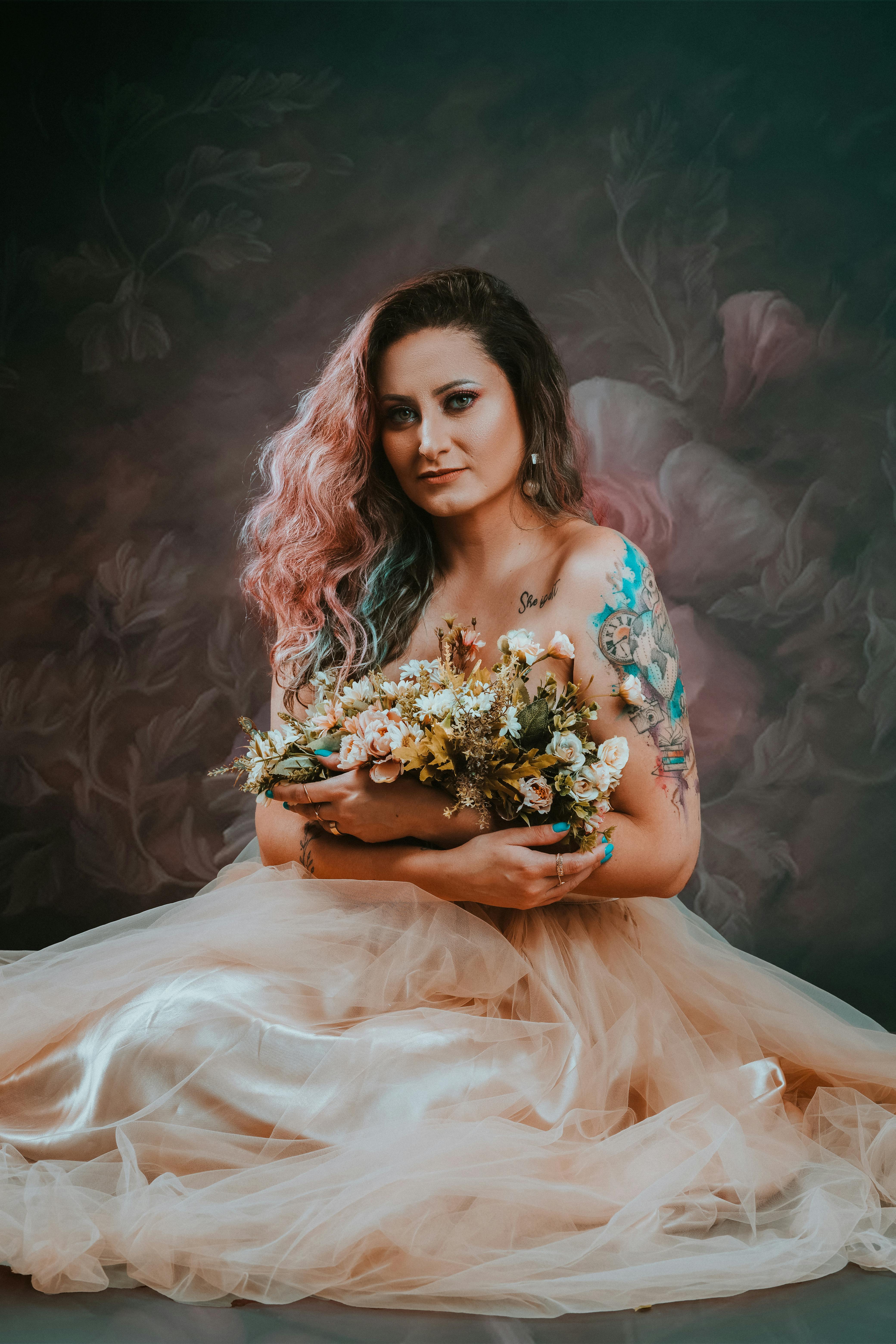 Portrait of a tattooed woman with pink hair holding a bouquet, wearing a flowing tulle dress in a studio setting.