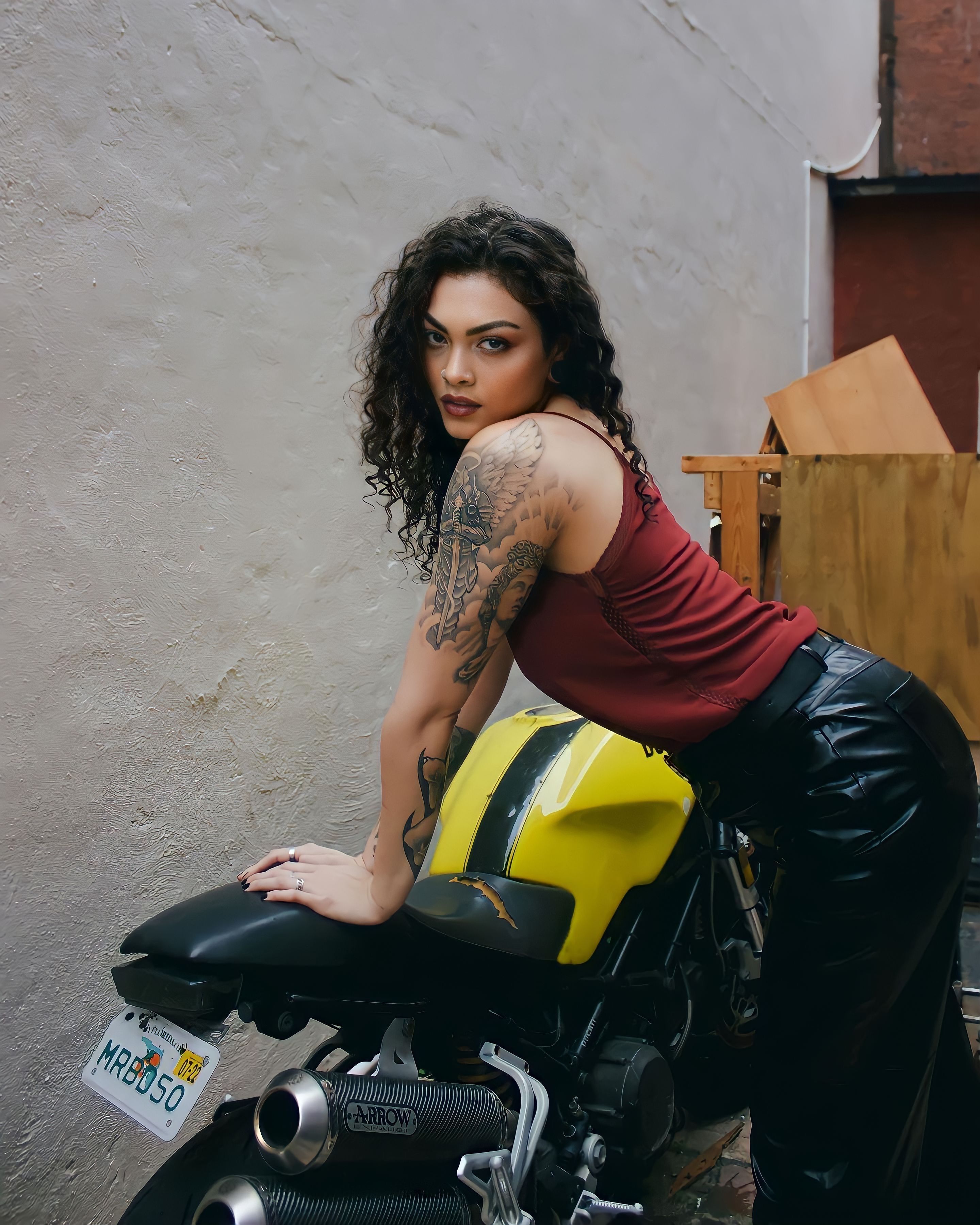 A fashionable woman with tattoos poses confidently with a motorcycle against an urban backdrop.