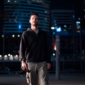 A man holding a camera stands confidently in a vibrant night cityscape.