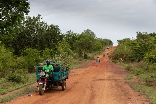 Dirt road in Dassa, Benin with motorcycles transporting goods through a lush, rural landscape.