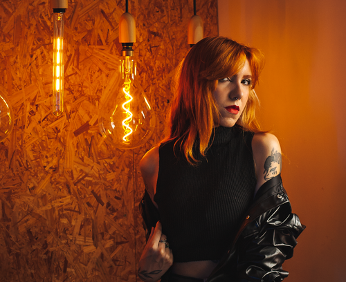 Free Tattooed woman with red lipstick poses beside glowing light bulbs indoors. Stock Photo