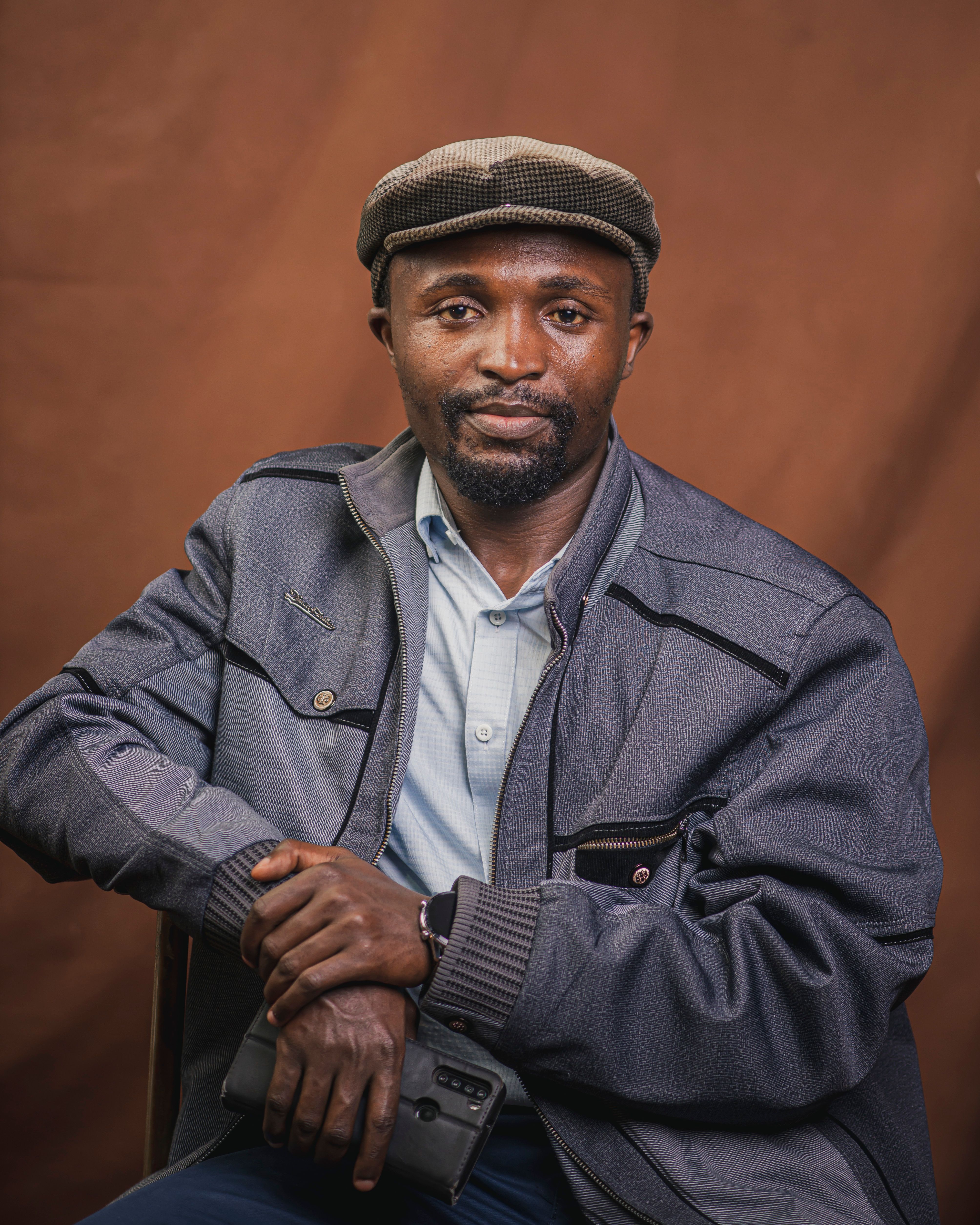 A fashionable man in a hat and jacket posing against a brown backdrop.