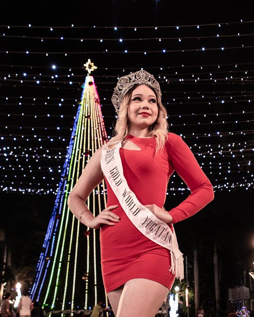 Woman in Crown and Red Dress Posing with Illuminated Christmas Tree behind
