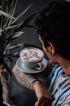 Tattooed man holding coffee cup with intricate latte art design, indoors setting.