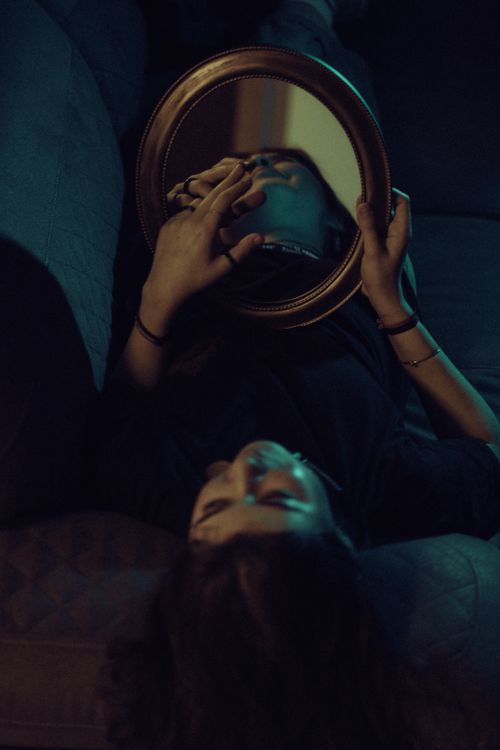 Free Moody indoor portrait showing a woman lying on a sofa, holding a mirror reflecting her face. Stock Photo