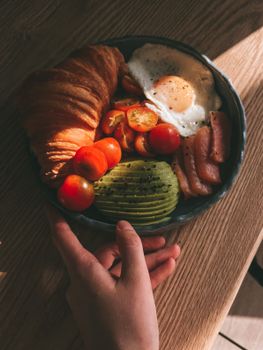 Appetizing breakfast bowl featuring croissant, avocado, tomatoes, egg, and salmon.