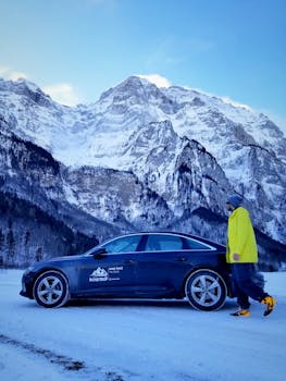 Man in yellow jacket walks by a black car against snowy mountain backdrop.