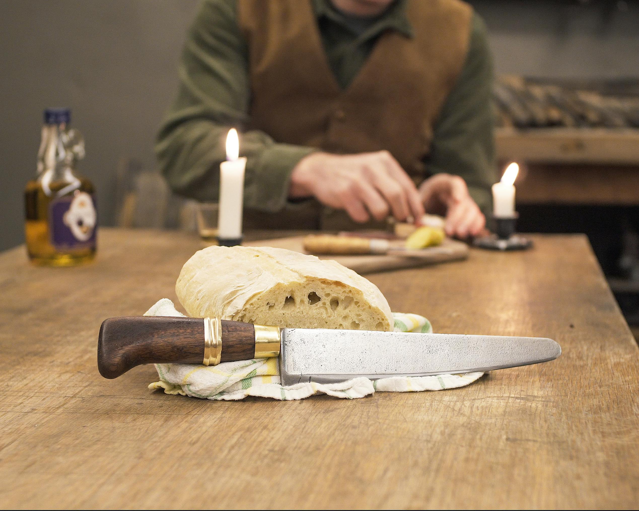 Rustic kitchen setup with bread and knife on wooden table, lit by candles.