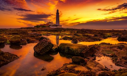 Free A stunning sunset over a rocky coast with a lighthouse and vibrant reflections. Stock Photo