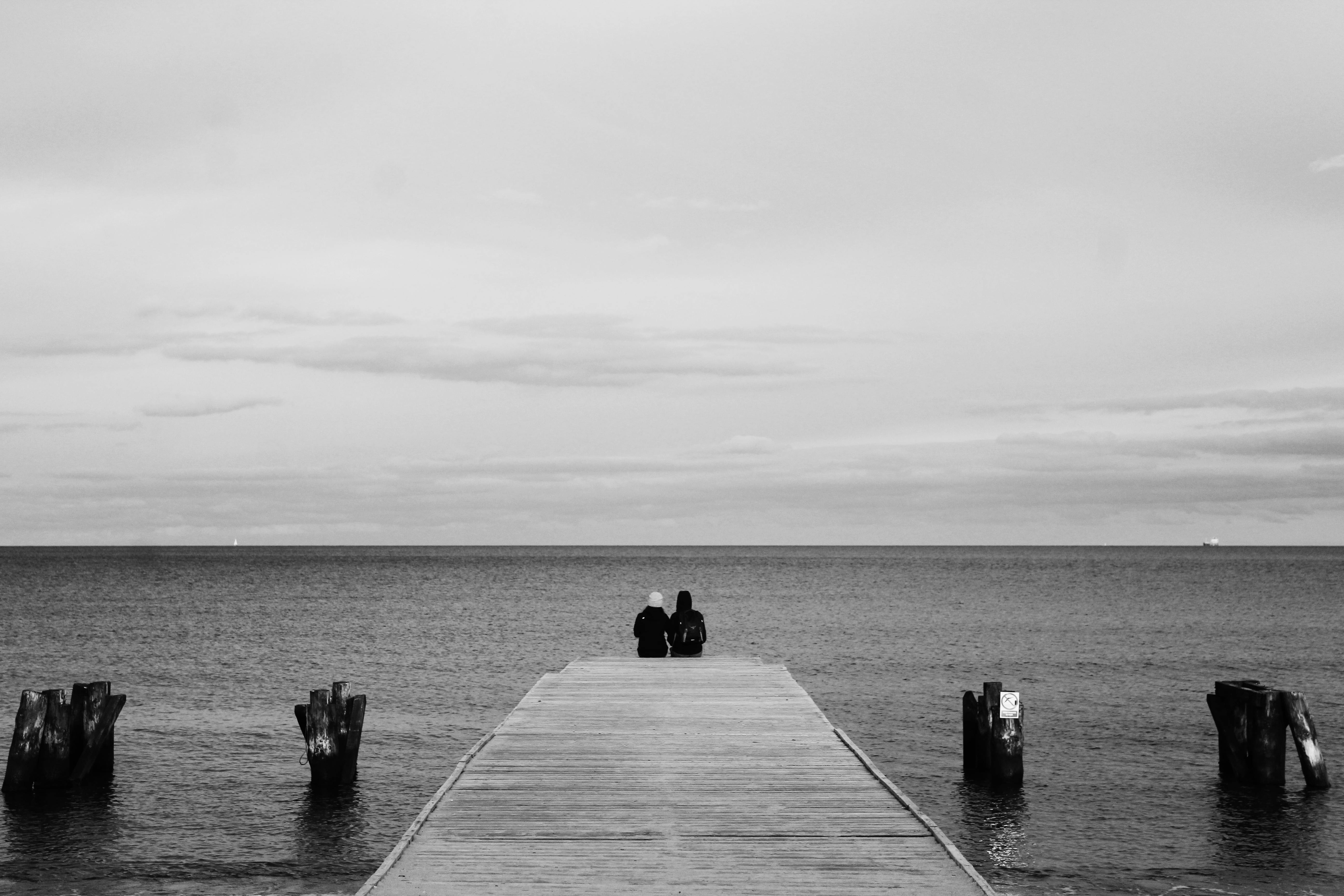 grayscale photography of couple sitting on wood dock