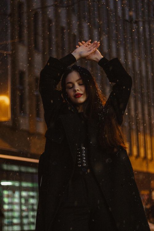 Free A fashionable woman in a black coat stands confidently at night with arms raised. Stock Photo