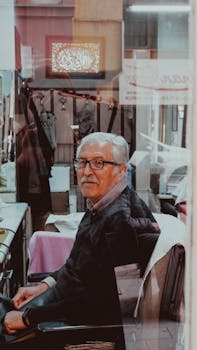Portrait of an elderly man with eyeglasses sitting in a barber shop seen through a window.