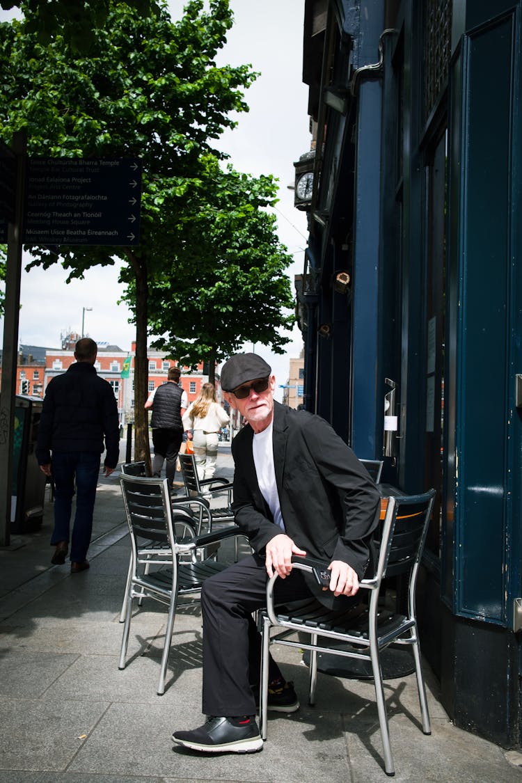 Elderly Man Sitting On A Chair In Front Of A Cafe In City 