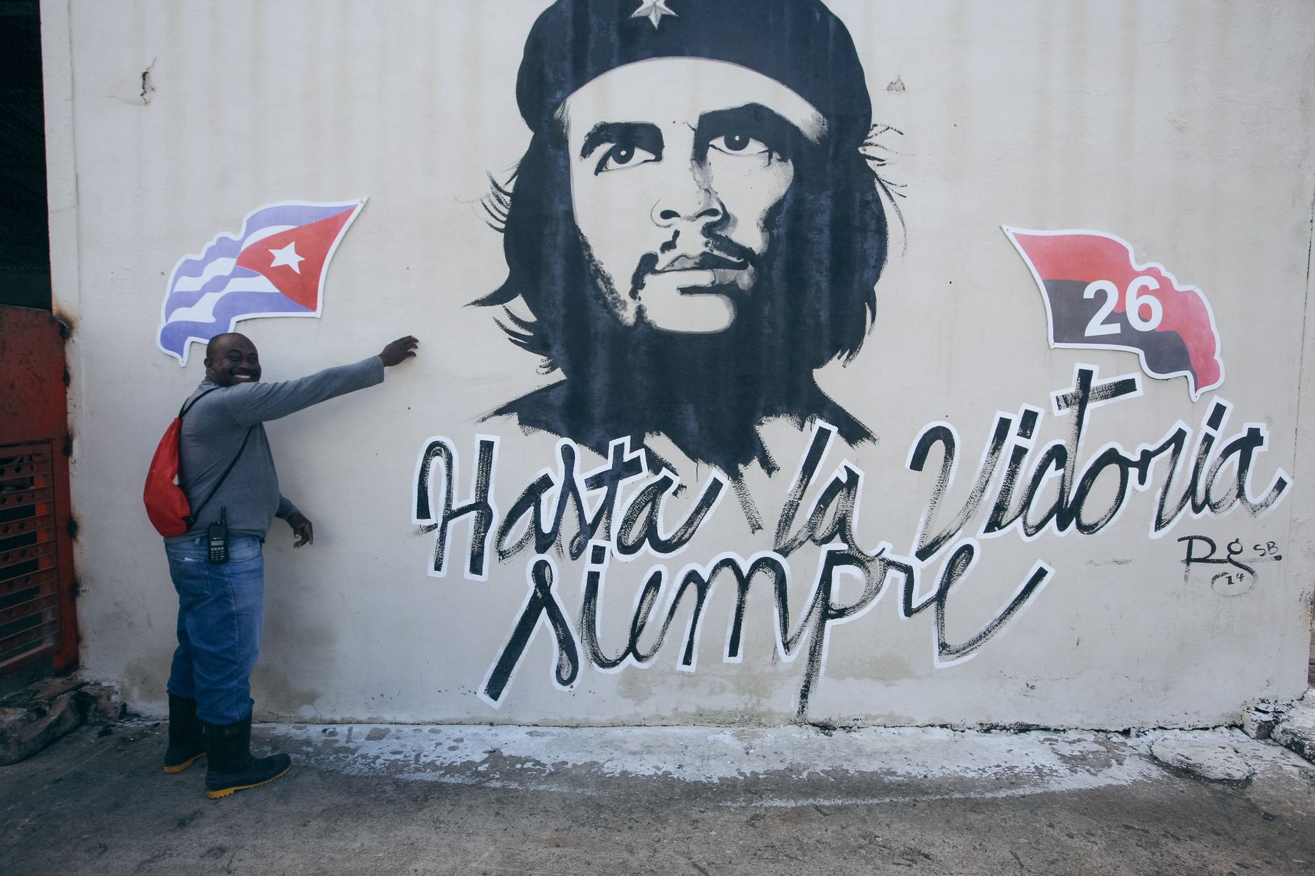 Man standing beside Che Guevara mural featuring Cuban flag and symbol of the 26th of July Movement.
