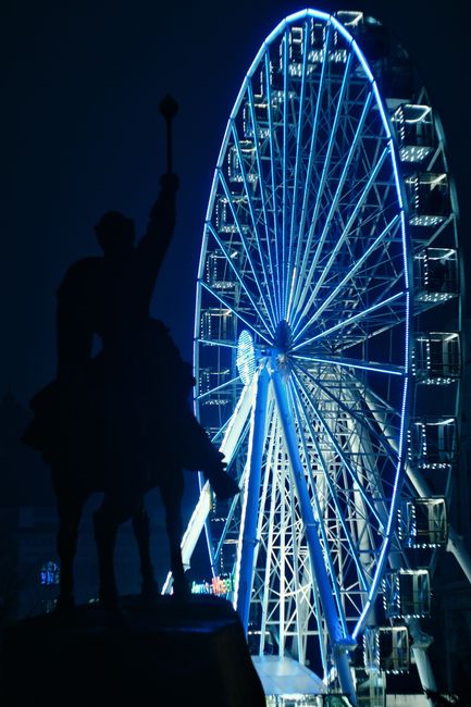 A striking image of an illuminated Ferris wheel at night, juxtaposed with a monument silhouette in Kyiv, Ukraine.