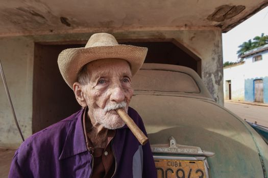 Senior man with hat smoking a cigar near a vintage car in Cuba.