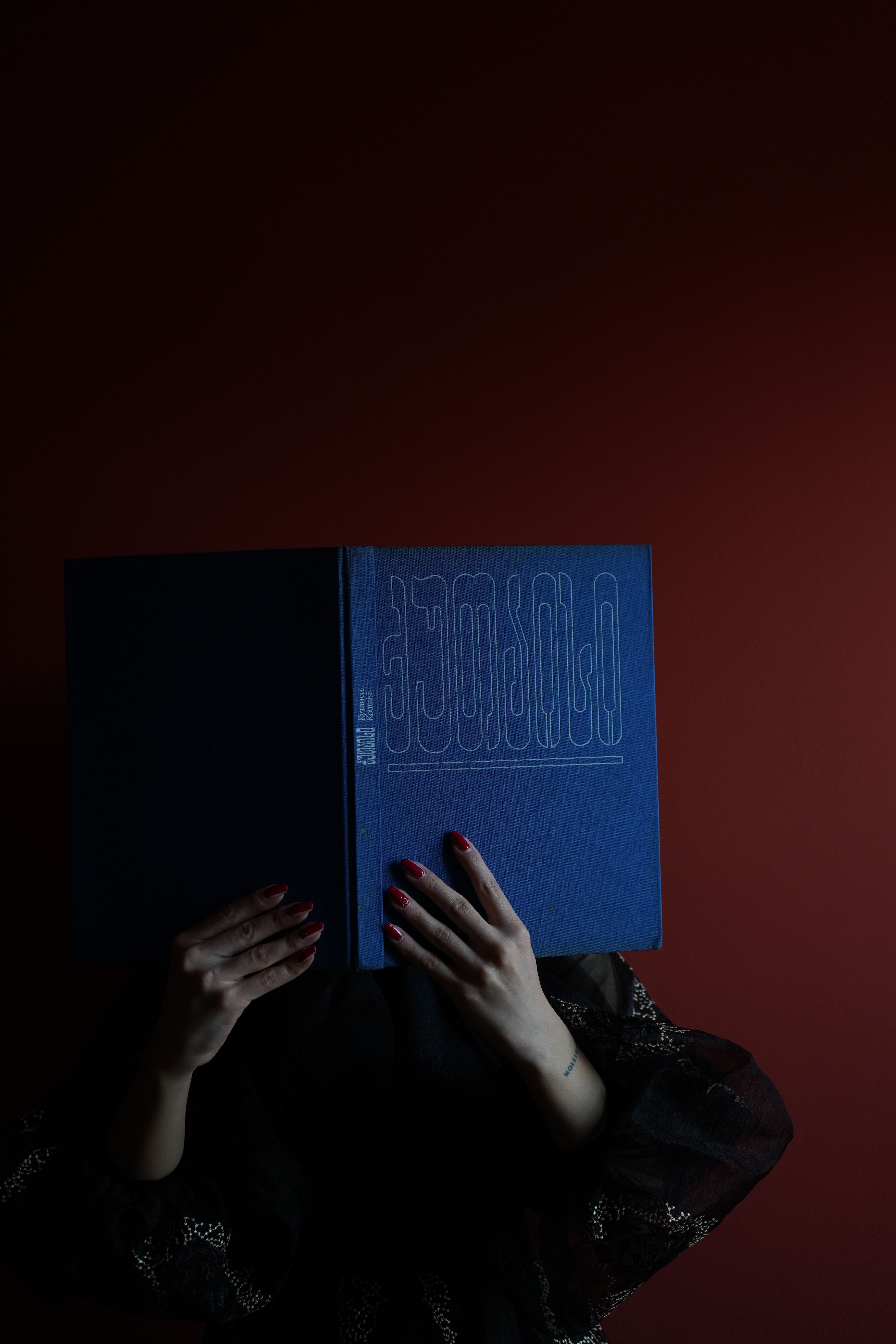Unrecognizable person holding a blue book, wearing a black blouse, with red nails, against a brown background.