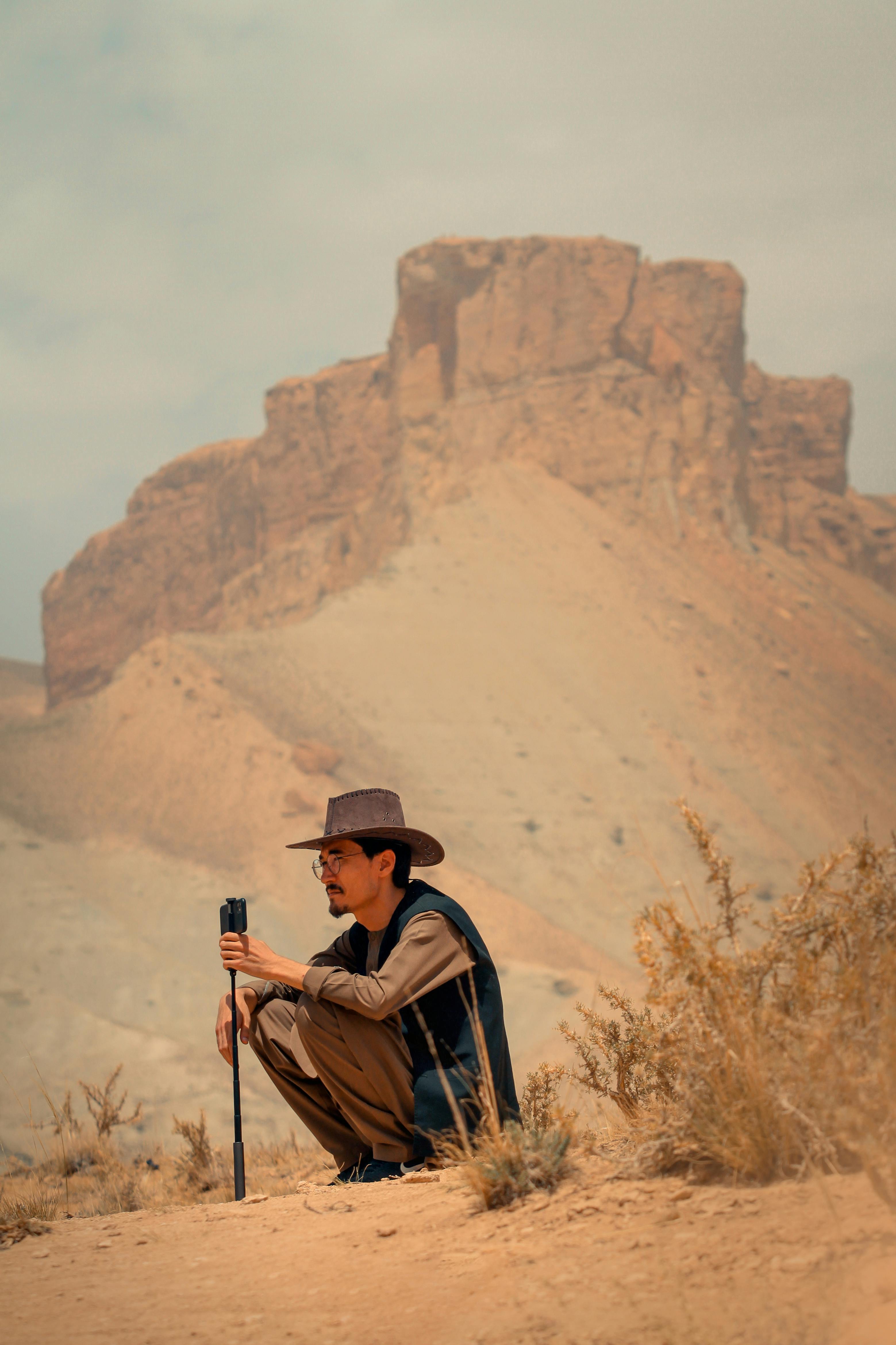 Man with hat and vest sitting in Bamyan's desert landscape.