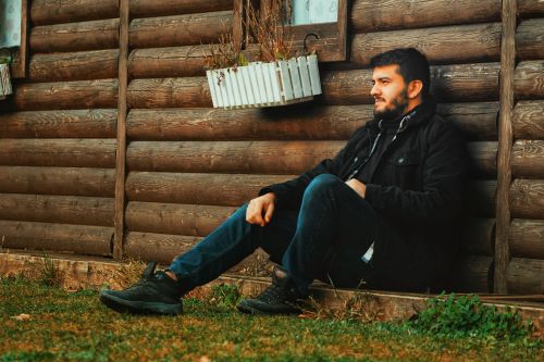 Free Stylish man in black attire sits against a rustic wooden wall, exuding a modern yet relaxed vibe. Stock Photo