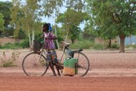 A young girl pushing a bicycle with containers in a rural field in Burkina Faso.