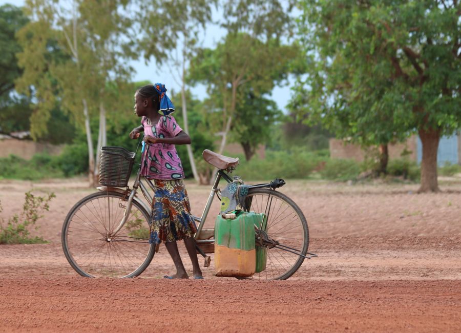 A young girl pushing a bicycle with containers in a rural field in Burkina Faso.