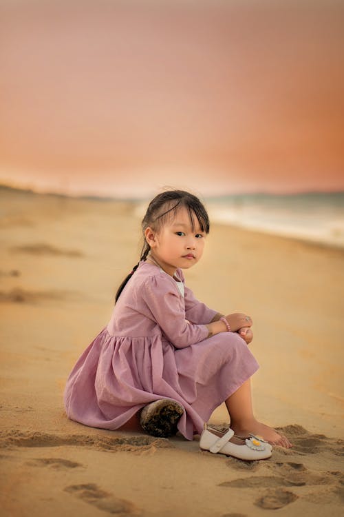 Little Girl in Purple Dress Sitting on Beach Sand