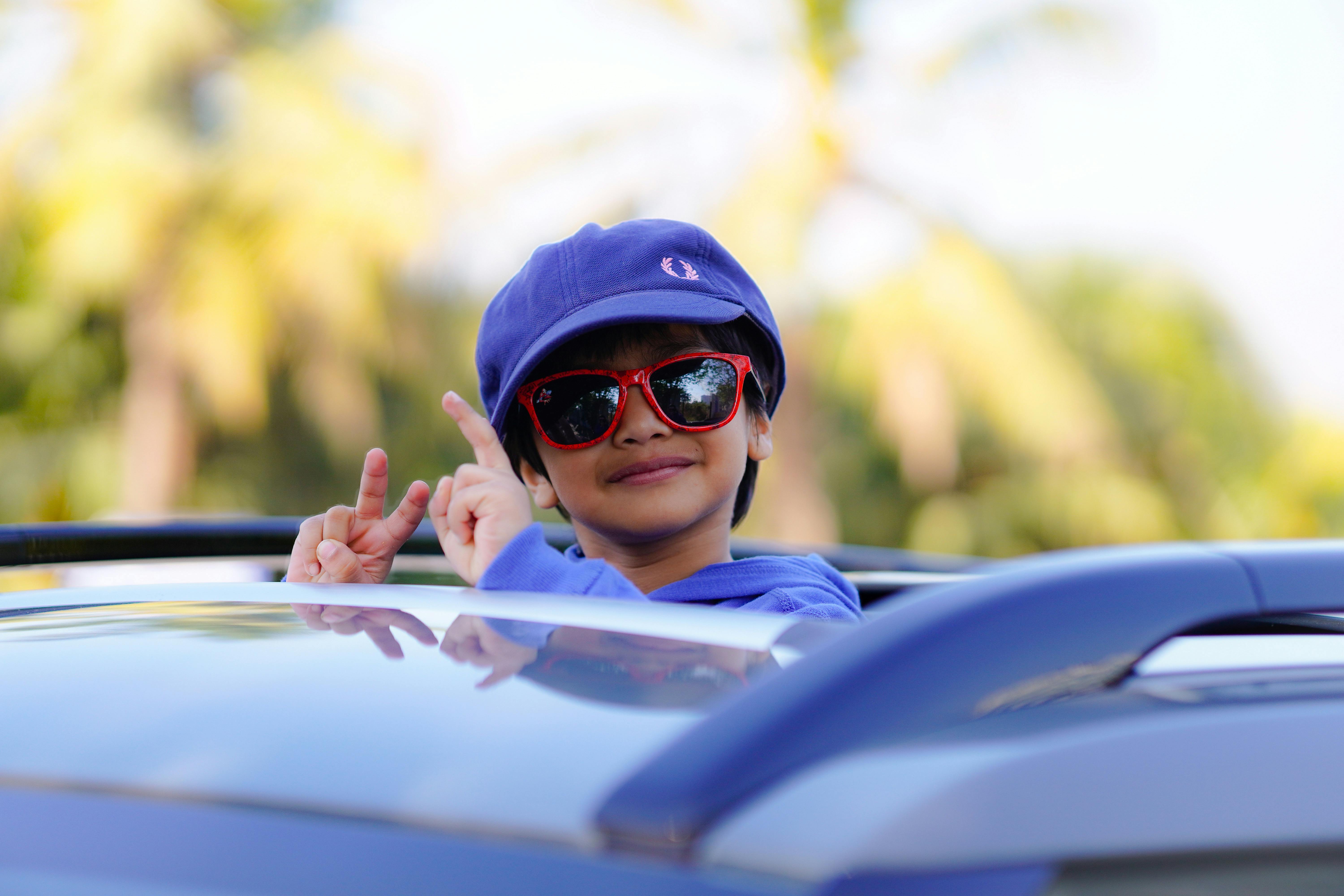 Cheerful child in blue cap and sunglasses smiling from a convertible car.
