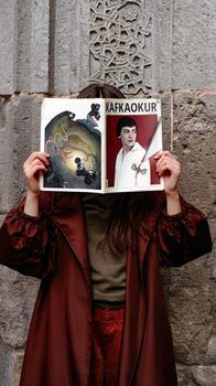 A woman in a brown coat holding a magazine with a Kafka feature against a stone facade.