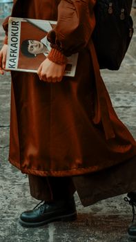 A stylish woman walking outdoors holding a magazine, dressed in a long brown coat and boots.