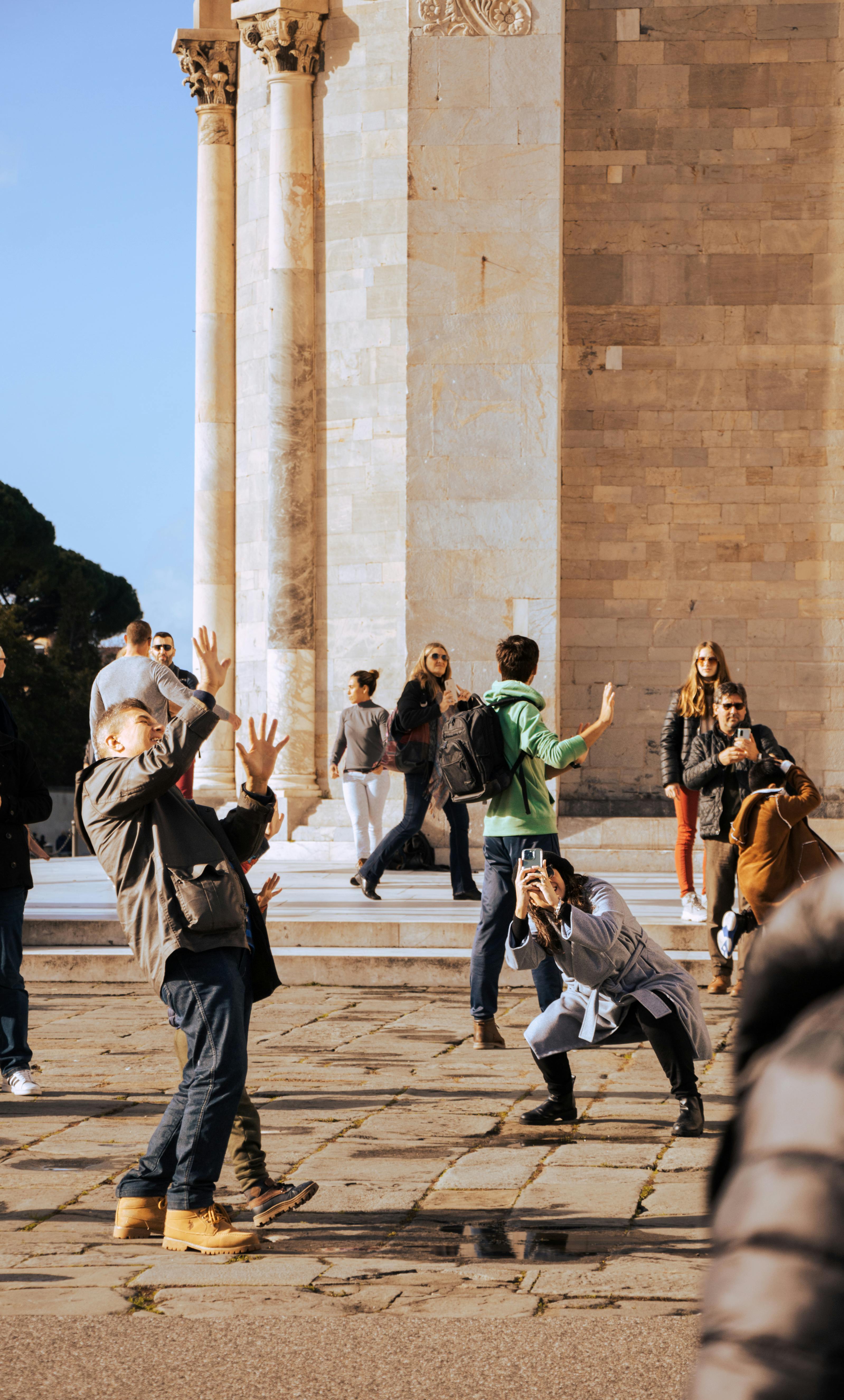 A lively scene of tourists posing and photographing near Pisa Cathedral on a sunny day.