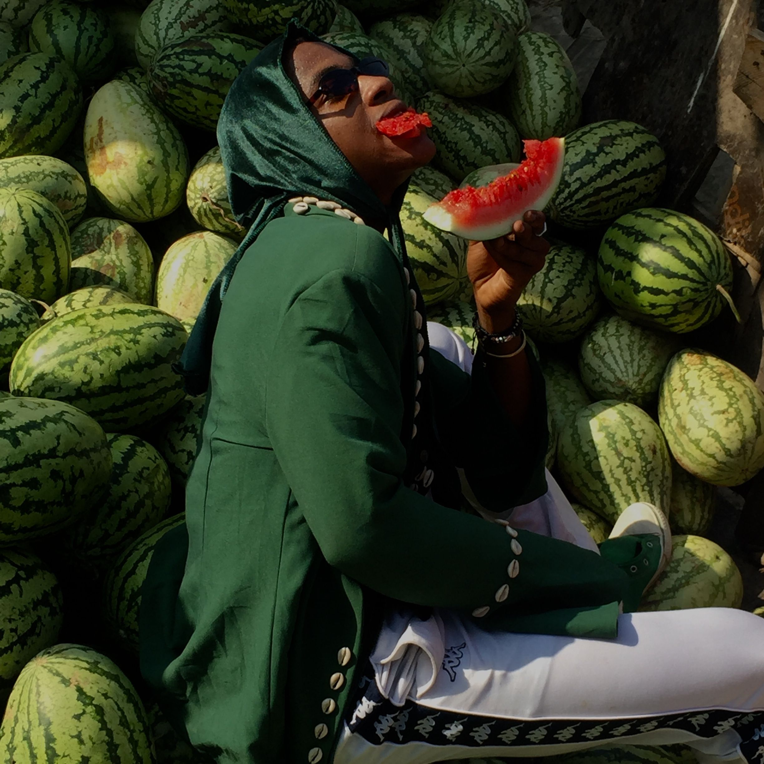 A fashionable woman in a green outfit enjoys watermelon while sitting among fresh melons outdoors.