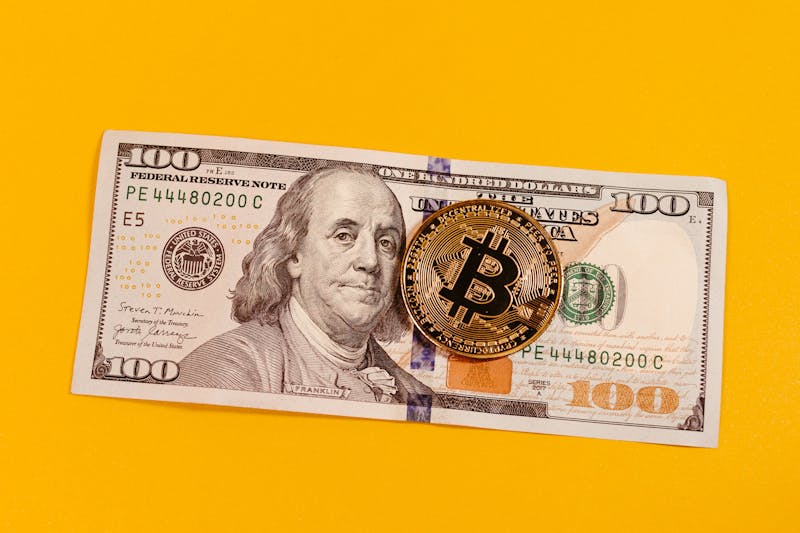 Gold bitcoin placed on a 100 US dollar bill over a bright yellow backdrop, symbolizing modern digital currency.