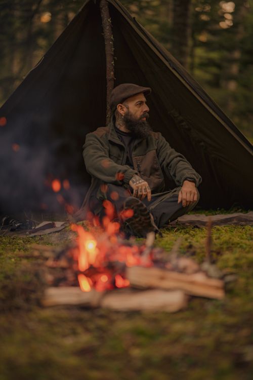 Free A bearded man sits by a campfire near a tent, enjoying nature. Stock Photo